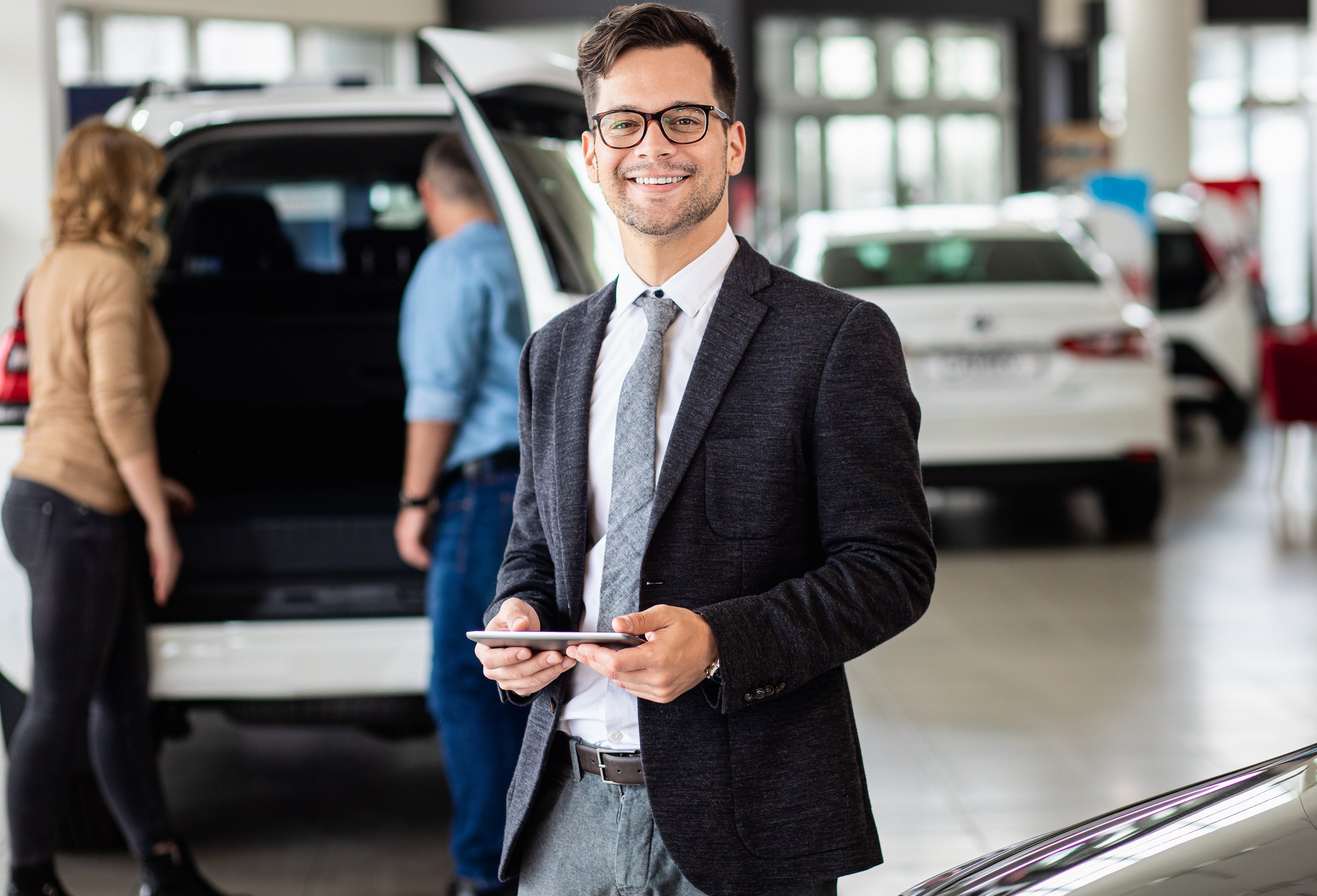 Car dealer holding a tablet in a showroom with vehicles behind him.