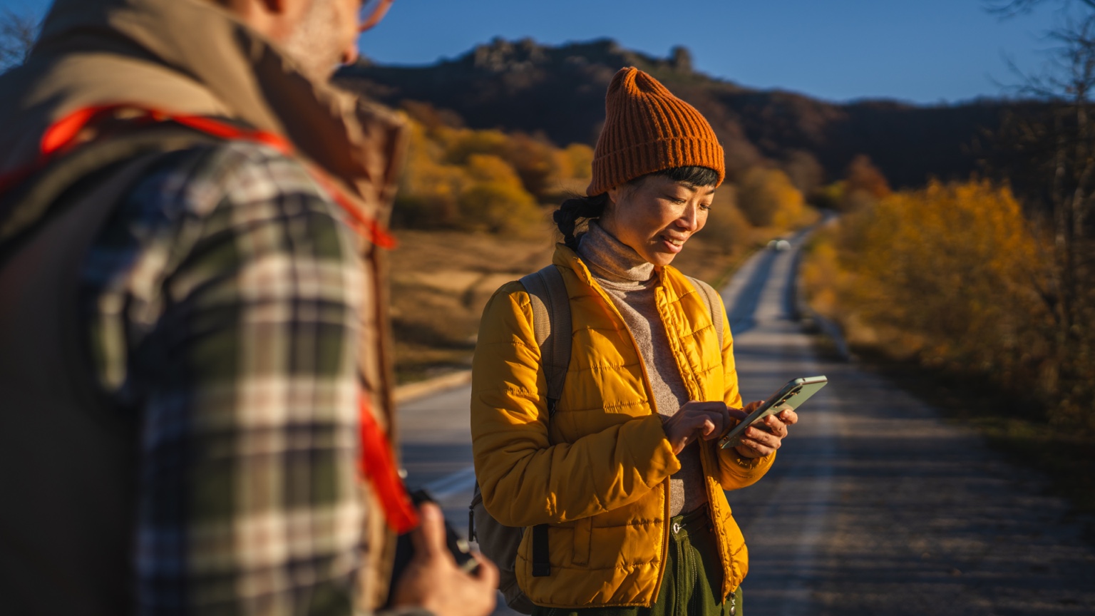 Woman in a yellow jacket checking her phone during an autumn hike with a companion
