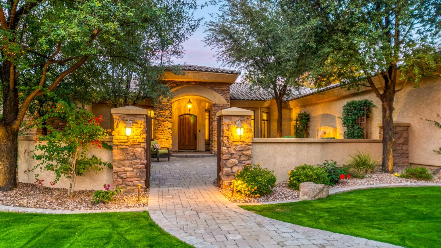 Luxury home with stone entry, mature trees, and warm exterior lighting