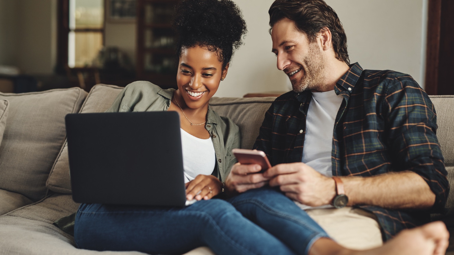 Couple using a laptop and phone together while seated on a sofa