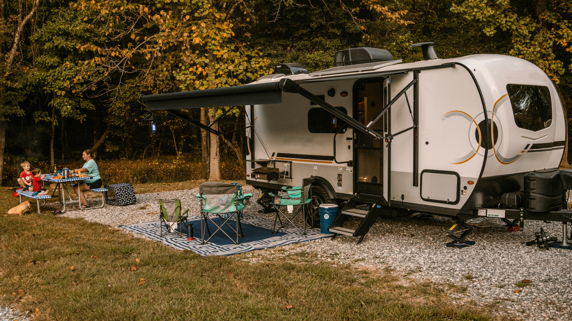 Family enjoying a picnic beside their travel trailer at a wooded campsite