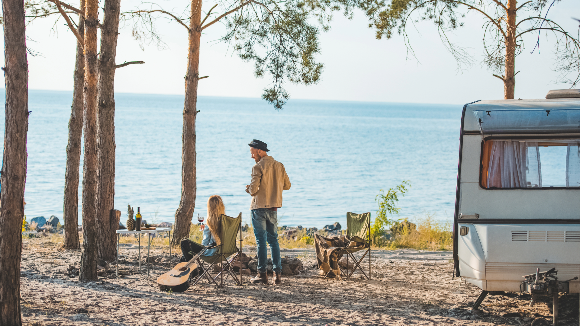 Couple relaxing with a guitar beside a travel trailer at a waterfront campsite